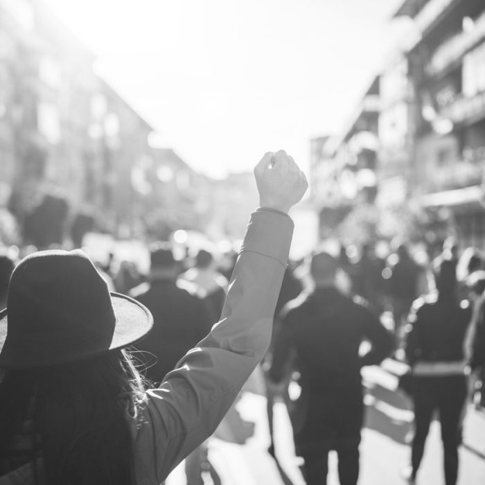 A young person raises their fist in a crowded street of protestors lit by the sun